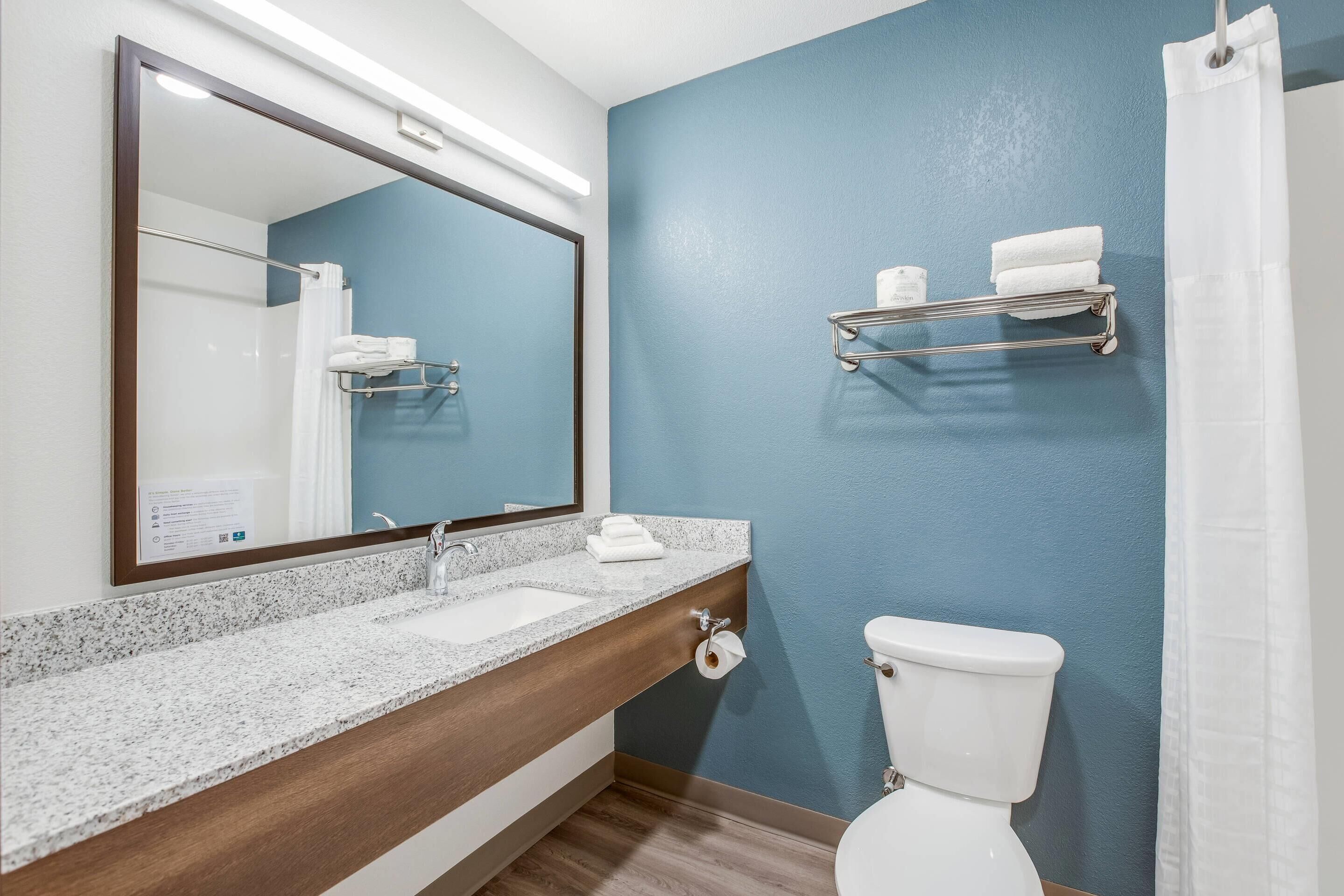 A bathroom with a granite countertop, a wooden cabinet, a white toilet, a blue wall, a mirror, and a towel rack.