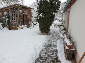 A snow-covered pathway leading to a wooden structure with a chimney, surrounded by snow-laden trees and a white building.