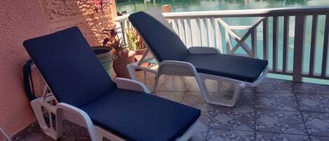 A balcony with two lounge chairs, a potted plant, and a view of a marina with boats and buildings.
