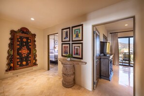 A hotel hallway with a decorative wooden panel, a stone pillar, framed pictures, and a balcony with a view.