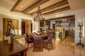 A spacious dining area with a round table and chairs, a chandelier, and a large painting on the wall.