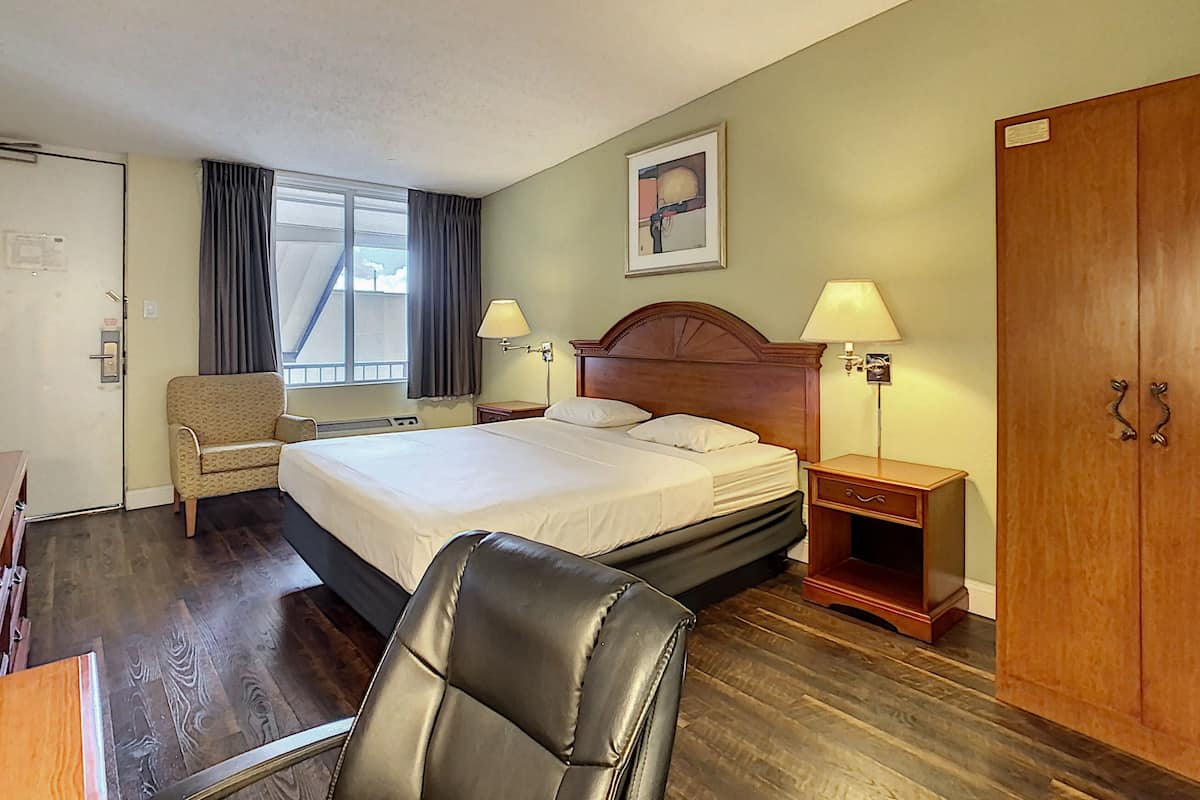 A hotel room with a large bed, a wooden wardrobe, a desk with a chair, and a window with curtains.