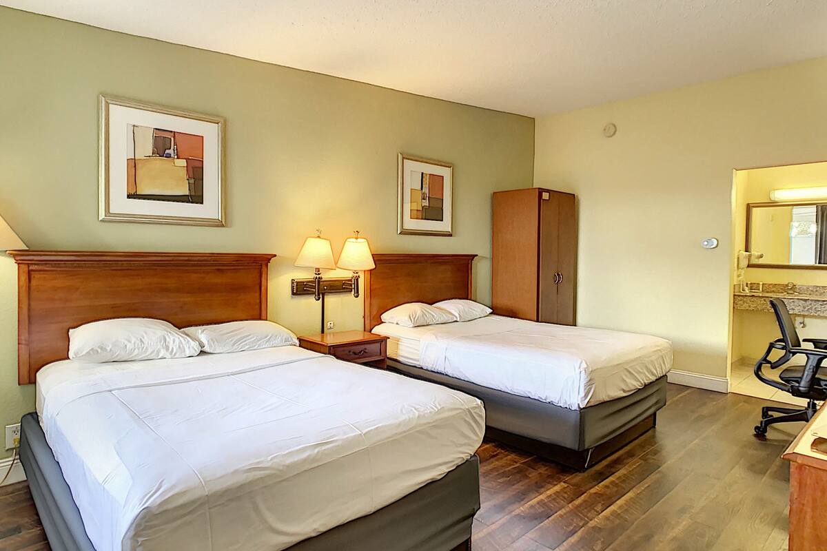 A hotel room with two beds, a wooden wardrobe, a desk with a chair, and two wall-mounted lamps.