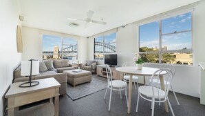 Ein modernes Wohnzimmer mit Sofa, Hocker, Fernseher, Esstisch, Stühlen und großen Fenstern mit Blick auf eine Brücke und Gebäude.