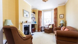 A living room with brown sofas, a fireplace, a bookshelf, and a window with curtains. - Large Spacious Family Home (sleeps 5), Parsons Green, LONDON (London)