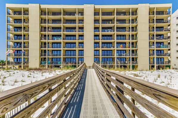 A multi-story hotel with balconies, a wooden boardwalk leading to the entrance, and a sandy beach in the background.