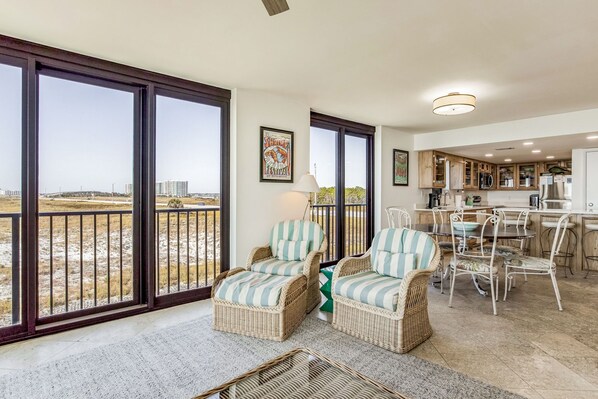 A spacious living area with wicker furniture, a dining table, and chairs. Large windows offer a view of the landscape.