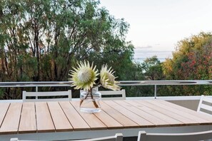 Une table en bois sur laquelle est posé un vase en verre transparent rempli de fleurs, le tout installé sur un balcon offrant une vue sur des arbres et une étendue d’eau.