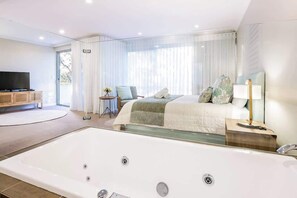 A modern hotel room with a large bathtub, a bed with a green blanket, a bedside table with a lamp, and a TV on a wooden cabinet. - Azure—Beachside Resort Escape for Groups (Marybrook)