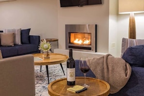 A cozy living room with a fireplace, a sofa, a coffee table with a bottle of wine and a glass, and a television mounted on the wall. - Azure—Beachside Resort Escape for Groups (Marybrook)