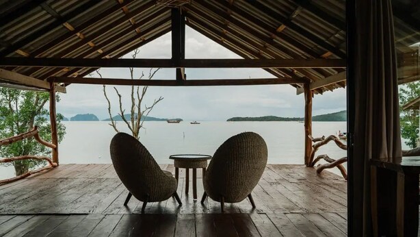Een houten terras met twee rieten stoelen gericht op een waterpartij, onder een rieten dakconstructie.