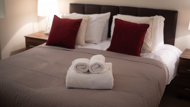 A neatly made bed with a brown bedspread, two red pillows, and two rolled white towels on the bed. - Newark House - 2 Bedroom Apartment (Maidenhall)