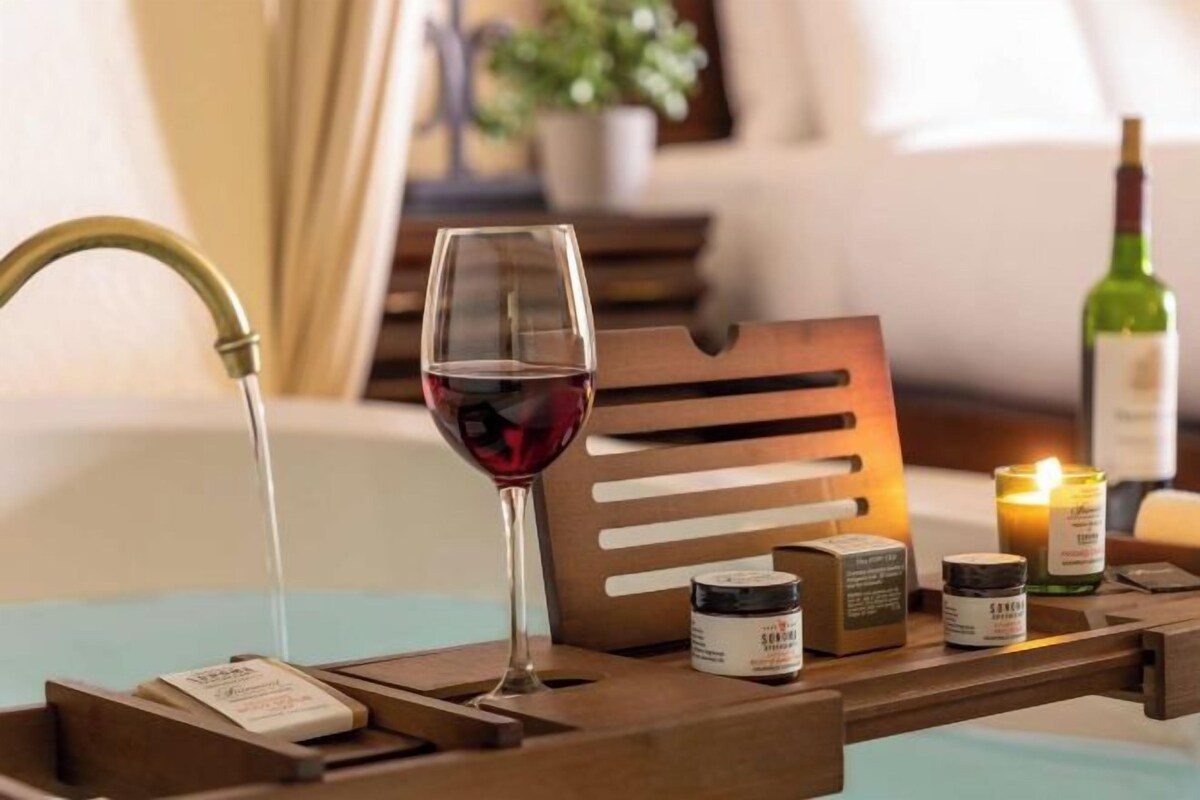 A wooden tray with a wine glass, candle, and small containers, likely for bath products, on a table.