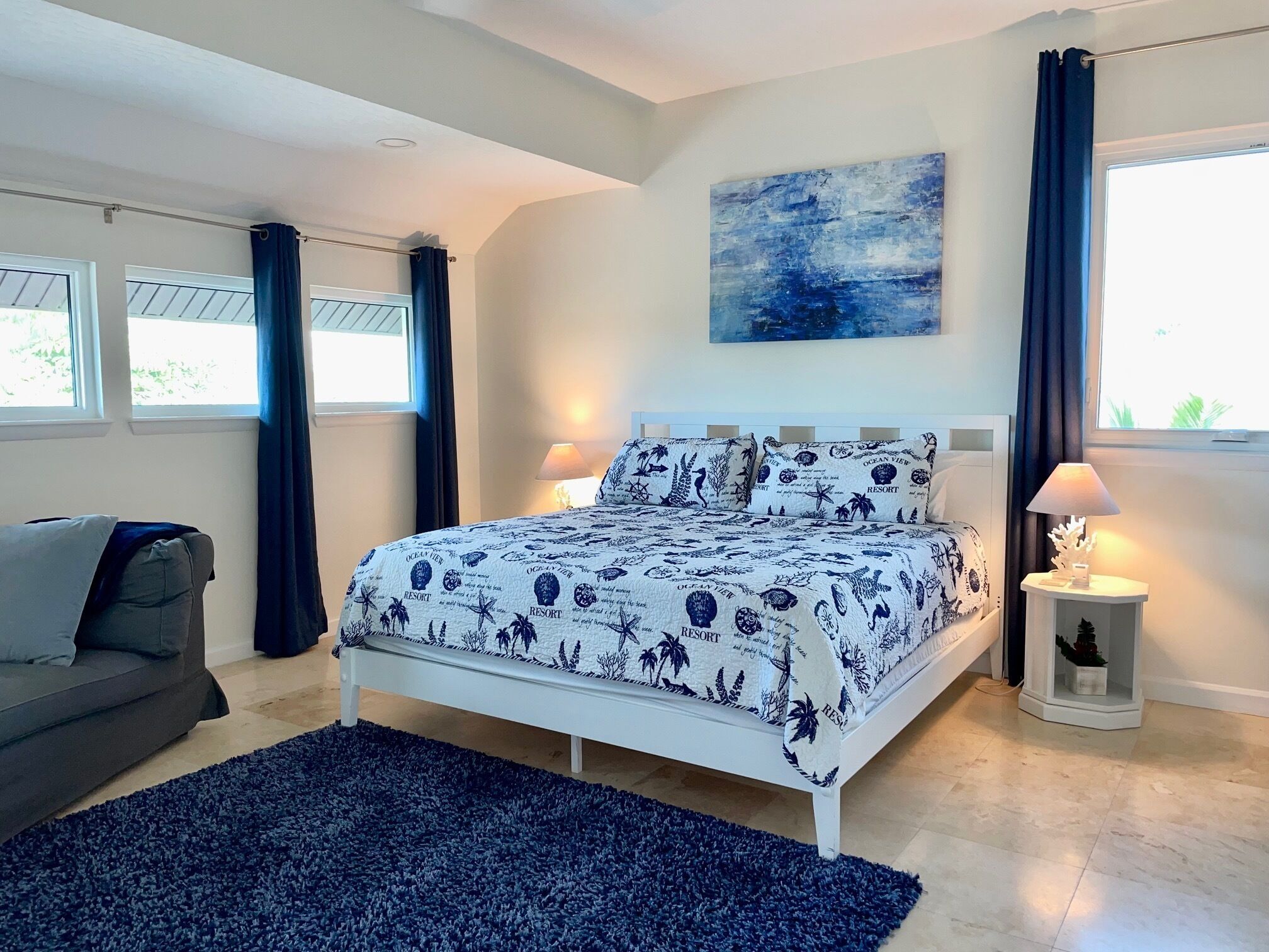 A bedroom with a bed, two bedside lamps, a nightstand, a window with curtains, a grey sofa, and a blue rug.