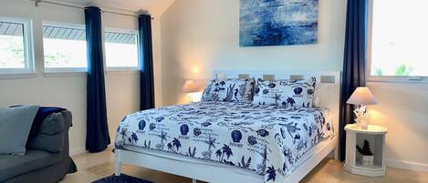 A bedroom with a bed, two bedside lamps, a nightstand, a window with curtains, a grey sofa, and a blue rug.