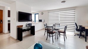 Una sala moderna con un televisor de pantalla plana, una mesa de comedor con sillas y un gran ventanal con persianas.