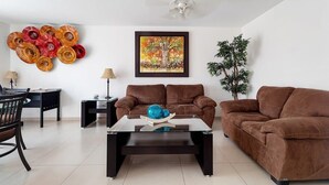 Una sala de estar con dos sofás marrones, una mesa de centro de vidrio, una mesa de comedor con sillas, una obra de arte en la pared y una planta en maceta.