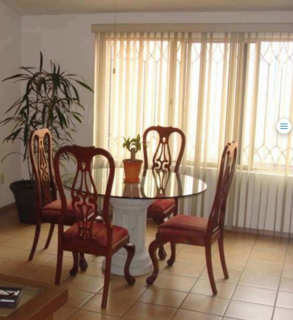 Een eetruimte met een ronde glazen tafel, omringd door vier houten stoelen met gedetailleerde rugleuningen. Op de tafel staat een potplant, en de kamer heeft een betegelde vloer en een raam met verticale zonwering.