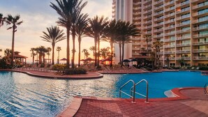 Zona de piscina en un resort, con palmeras, sillas de descanso y un edificio alto al fondo.