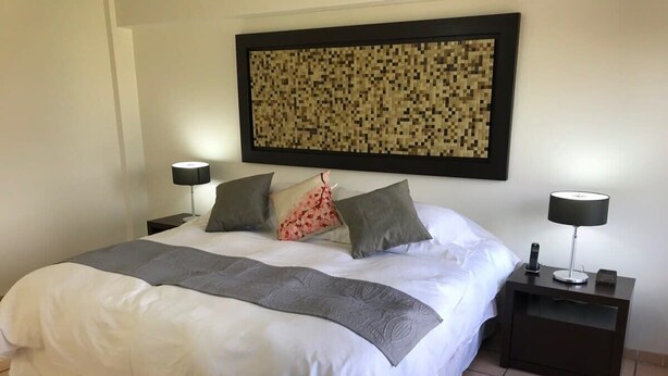 A neatly made bed with white bedding and a gray throw, flanked by two nightstands with lamps, in a room with a tiled floor and a large pixelated artwork on the wall. - +MS +Suite Perfecta +Centro +Av Zaragoza +S5 (Santiago de Querétaro)