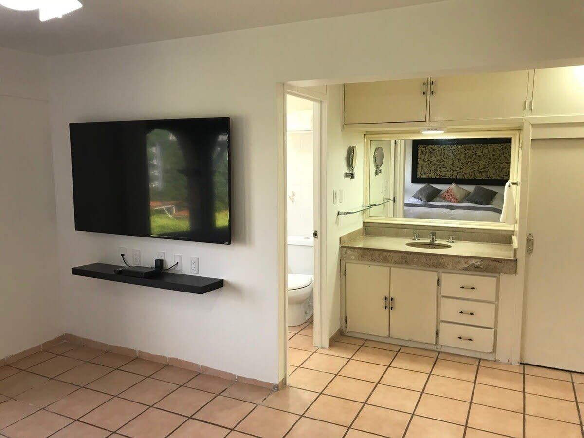 A small, clean room with a flat-screen TV mounted on the wall, a small shelf below it, and a kitchenette area with a sink and cabinets.