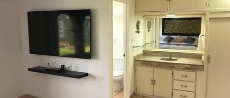 A small, clean room with a flat-screen TV mounted on the wall, a small shelf below it, and a kitchenette area with a sink and cabinets.