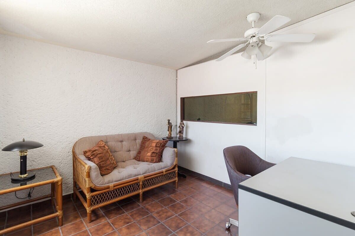 Una habitación de esquina con un sofá de mimbre, una mesita con una lámpara, un ventilador de techo y un armario blanco.