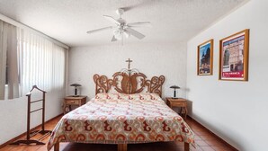 A bedroom with a bed, a ceiling fan, a cross on the wall, and two framed pictures.