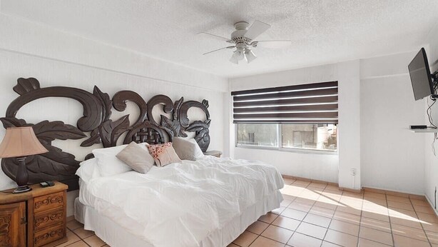 Une chambre avec un grand lit, un ventilateur de plafond, une fenêtre avec des stores et un téléviseur fixé au mur.