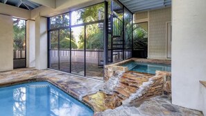 An indoor swimming pool with a stone design, large glass windows, and a view of greenery outside.