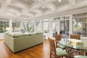 A spacious room with a green sofa, wicker chairs, a glass table, and large windows offering a view of the outdoors.