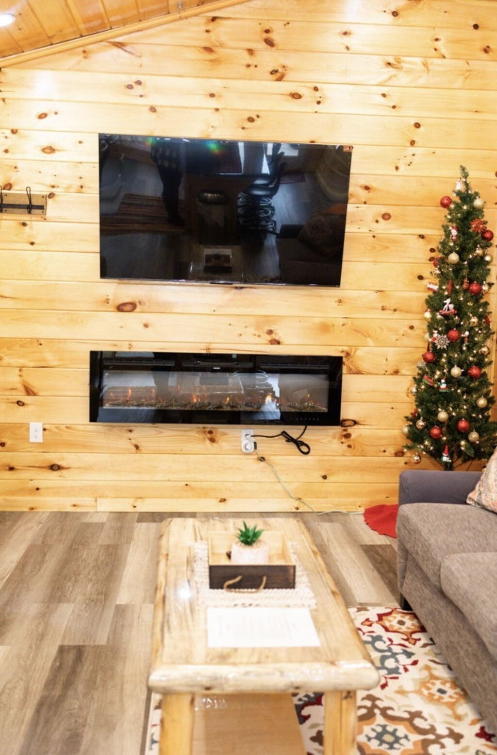 Una habitación con paneles de madera, un televisor de pantalla plana montado en la pared, una chimenea debajo, un pequeño árbol de Navidad y una alfombra estampada.
