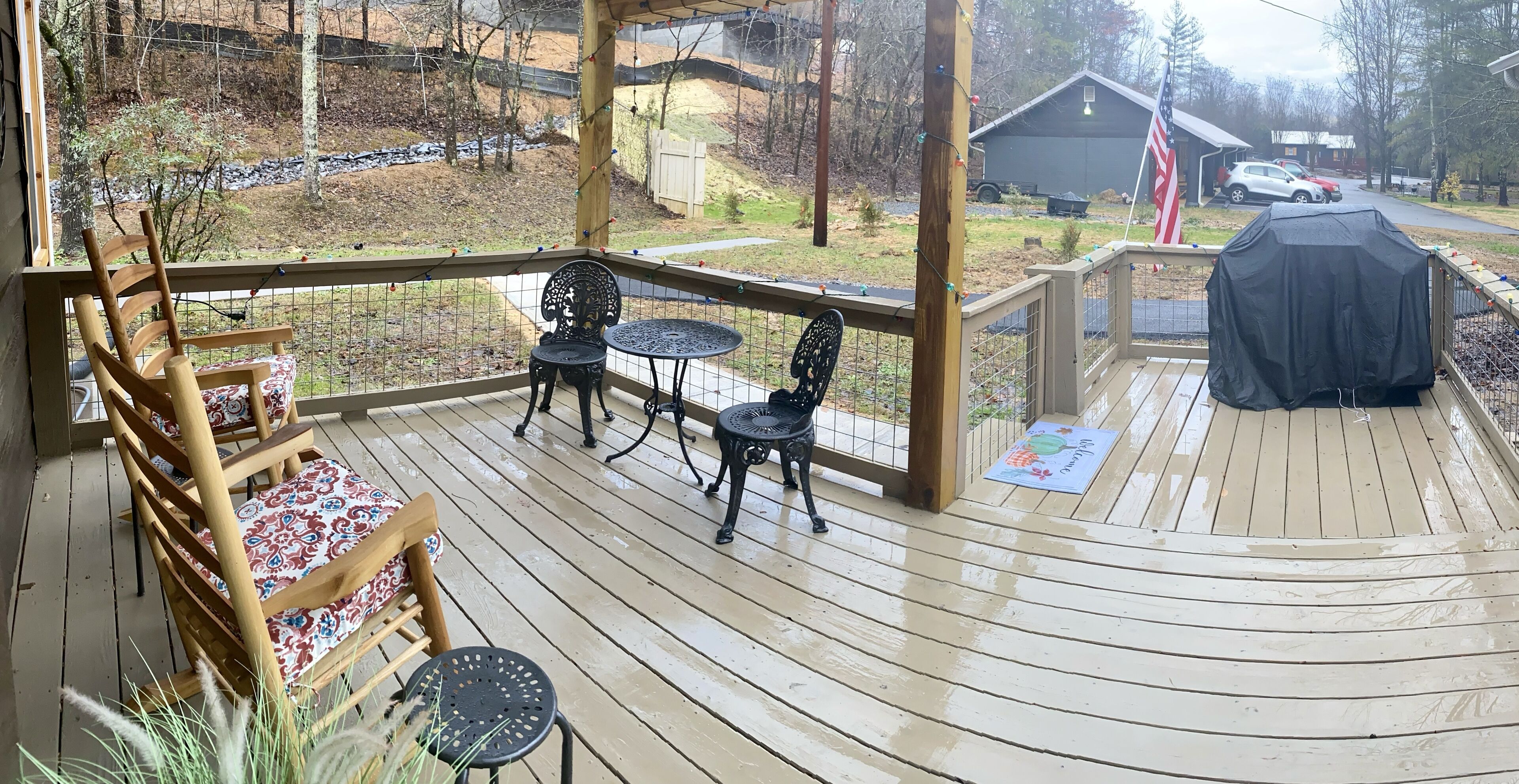 A wooden deck with outdoor seating, including chairs and tables, overlooking a yard with a covered car and a barn in the background.