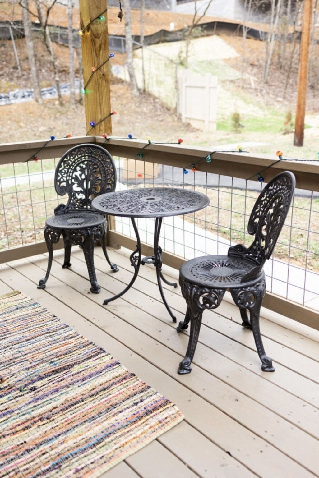 A wooden deck with two ornate metal chairs and a small round table, surrounded by a metal railing and string lights.