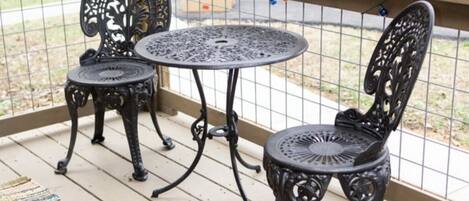 A wooden deck with two ornate metal chairs and a small round table, surrounded by a metal railing and string lights.