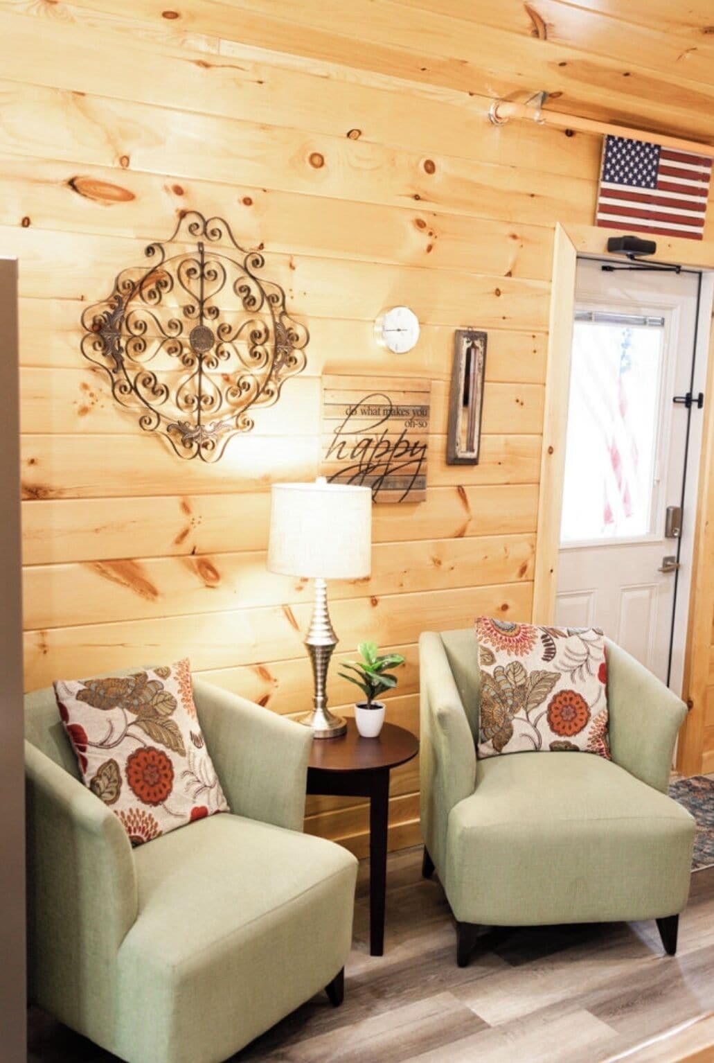 A cozy room with wooden walls, two green armchairs with floral cushions, a small table with a lamp, a potted plant, and a wall clock.