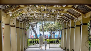 A covered walkway with a tiled floor, flanked by columns and plants, leading to a gated area with a view of palm trees and the sea.