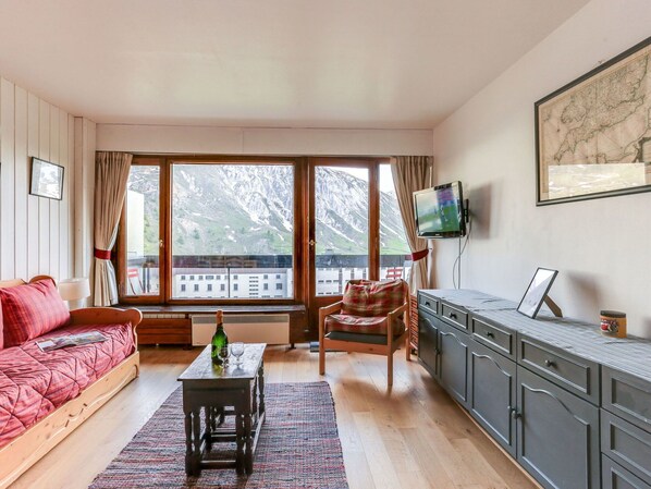 A cozy living room with a sofa, a chair, a coffee table, and a TV. Large windows offer a view of mountains and a building.