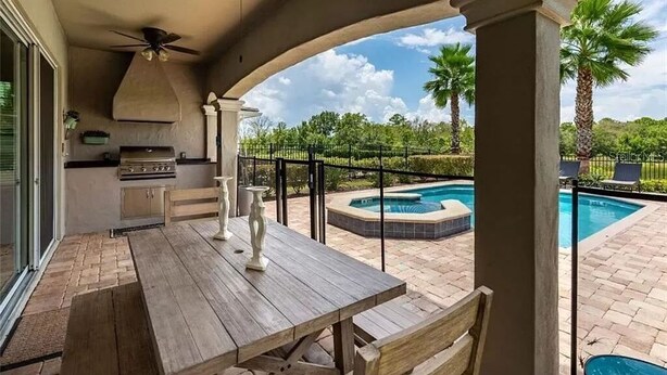 Eine Terrasse im Freien mit einem Holztisch, einem Deckenventilator und Blick auf einen Swimmingpool und Palmen.