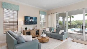Ein modernes Wohnzimmer mit einer Couch, einem Couchtisch und einem Fernseher. Eine Schiebetür führt zu einem Poolbereich.