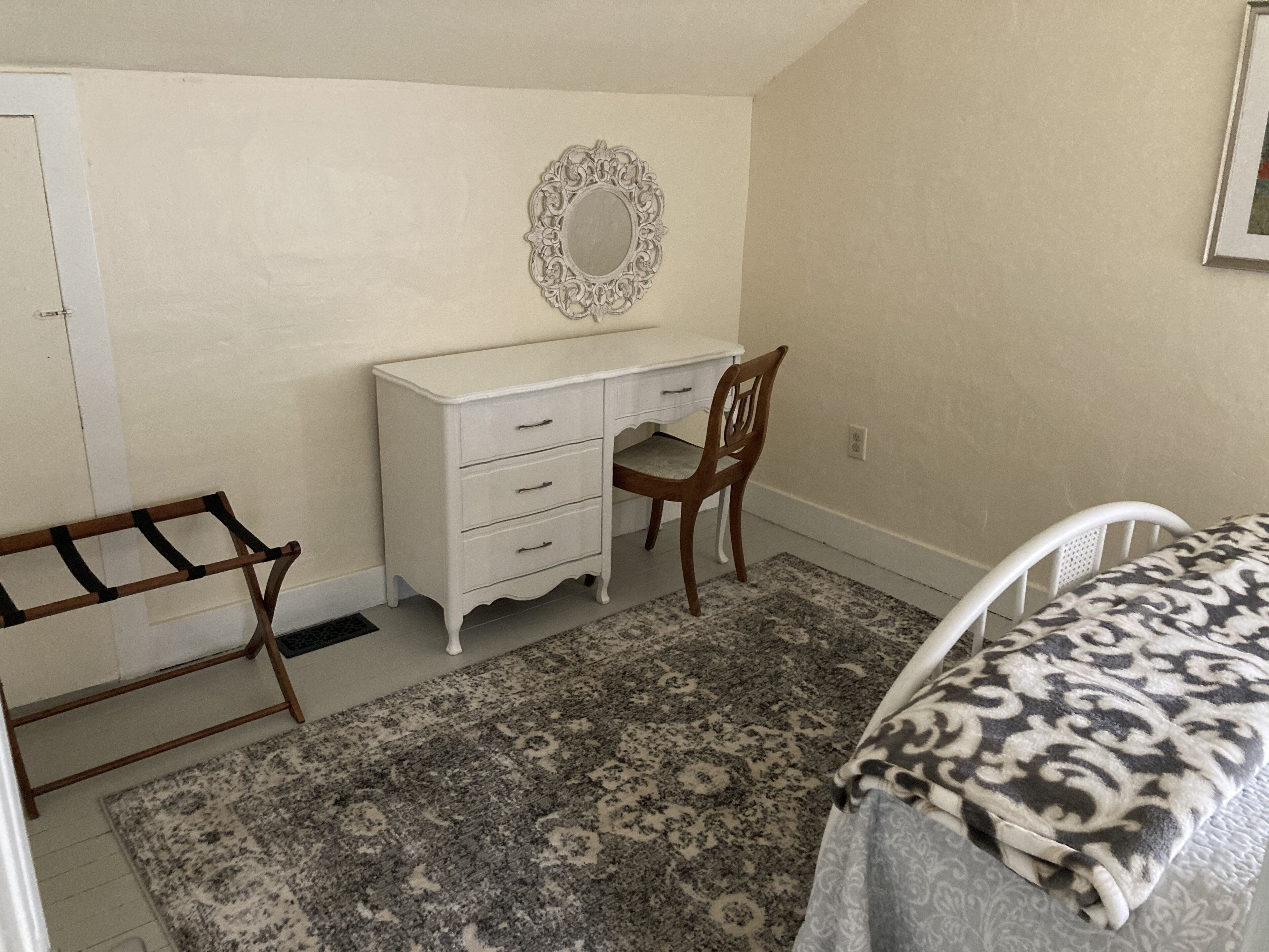 Um quarto com cama, escrivaninha com cadeira, cômoda e espelho na parede.