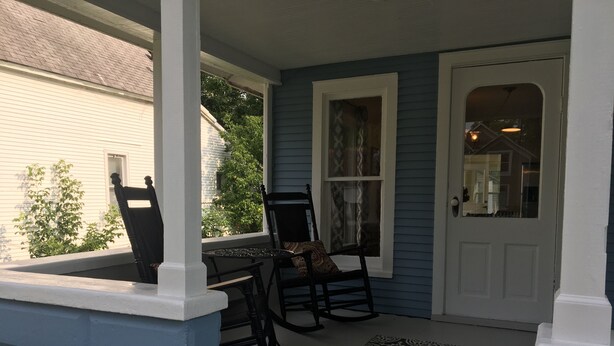 A porch with a rocking chair and a table, a patterned rug, and a white door.