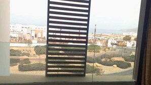 Ein Fenster mit horizontalen Jalousien, Blick auf eine Küstenlandschaft mit Gebäuden und Grünflächen.