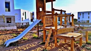 A wooden play structure with a slide and climbing elements, set on sandy ground with modern buildings in the background. - Residence Tamourrit Taghazout Bay 4747 (Taghazout)
