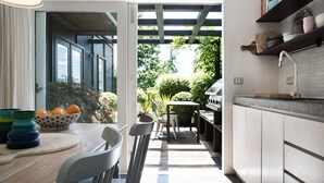 A modern kitchen with white cabinets, a dining table with chairs, and a view of a garden through an open door. - Harts Lane Stag and Millies- sleek design and pet friendly (Daylesford)