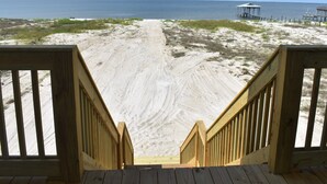 Una escalera de madera que conduce a una playa arenosa, con vista al océano y una construcción pequeña en la distancia.