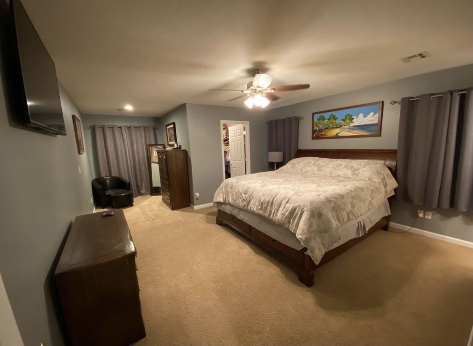 A bedroom with a bed, a dresser, a chair, a ceiling fan, and a painting on the wall.
