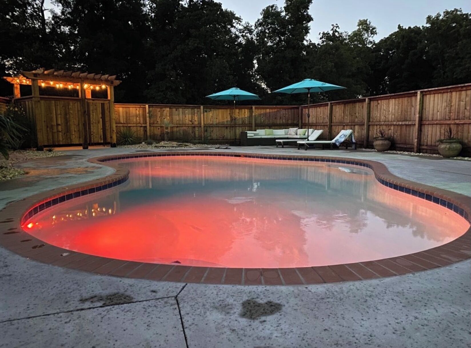 Piscina iluminada com luz vermelha, cercada por uma vedação de madeira, e área de descanso com guarda-sóis.