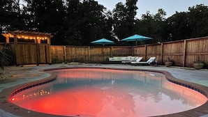 Une piscine éclairée de lumières rouges, entourée d’une clôture en bois, et un espace détente avec des parasols.