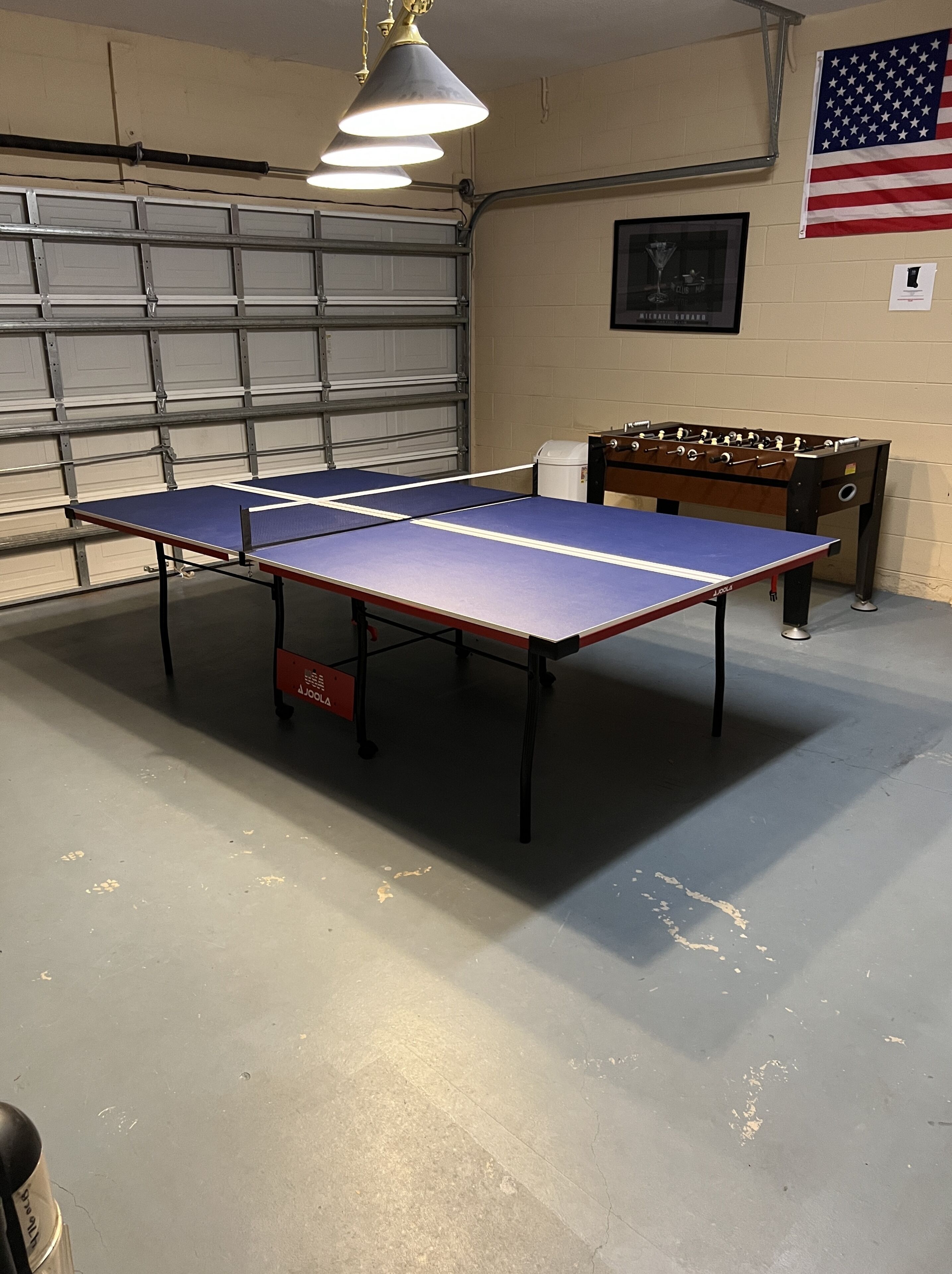 A ping pong table in a garage with a foosball table and an American flag on the wall.